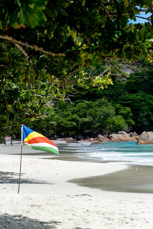 Seychelles flag stuck in the sand on the beach, behind it a restless ocean with large rocks lying in it.