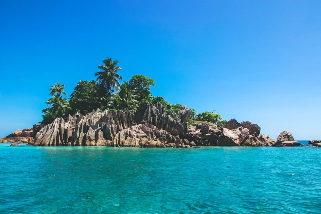 A beautiful island covered with rocks on the side, with palm trees on top and a wonderful ocean around.